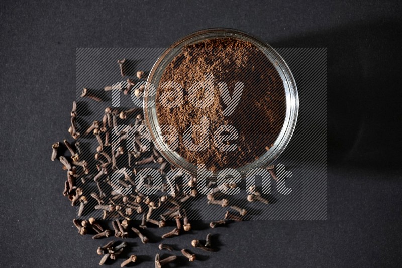A glass bowl full of cloves powder with cloves grains spread on black flooring