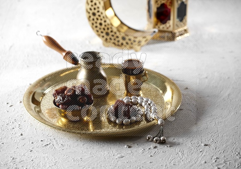 Dates in a metal bowl with coffee and prayer beads on a tray beside lanterns in a light setup