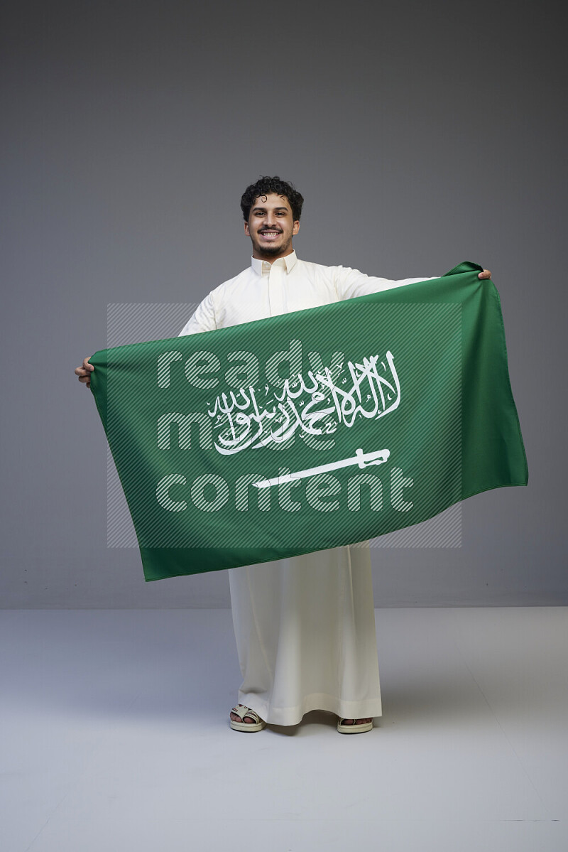 A Saudi man standing wearing thob holding big Saudi flag on gray background