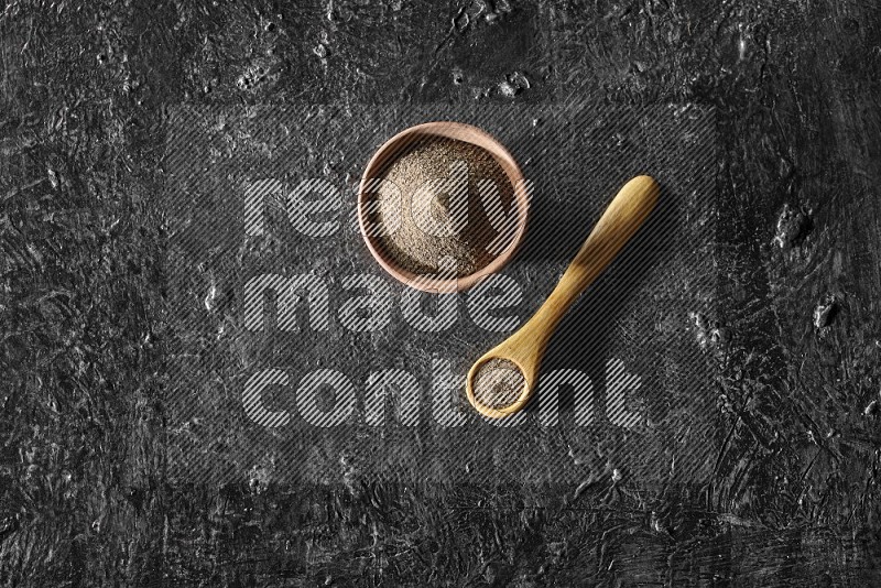 A wooden bowl and wooden spoon full of black pepper powder on a textured black flooring