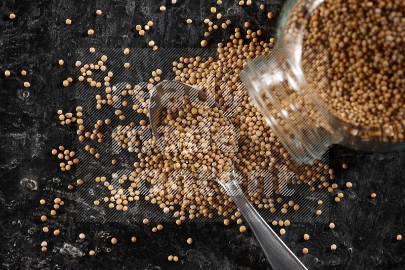 A glass spice jar and a metal spoon full of mustard seeds and jar is flipped with fallen seeds on a textured black flooring