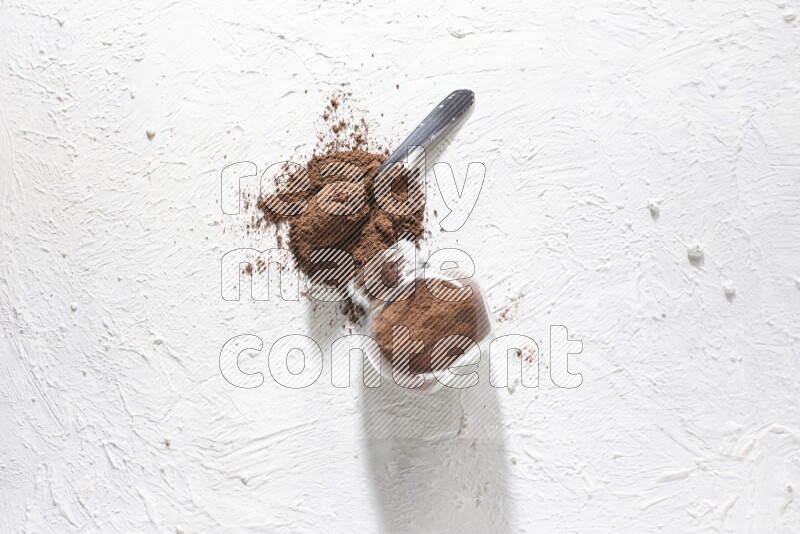 A flipped glass spice jar and a metal spoon full of cloves powder and powder came out of the jar on textured white flooring