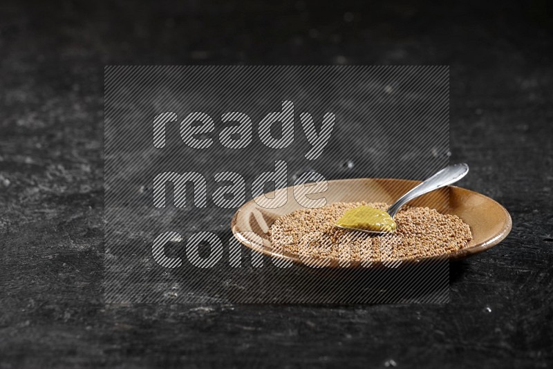 A beige pottery plate filled with mustard seeds with a metal spoon full of mustard paste in it on a textured black flooring