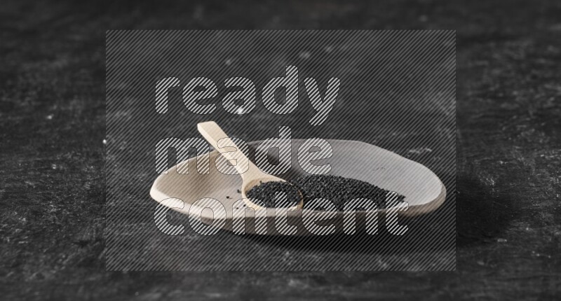 A multicolored pottery plate full of black seeds and wooden spoon full of seeds on a textured black flooring
