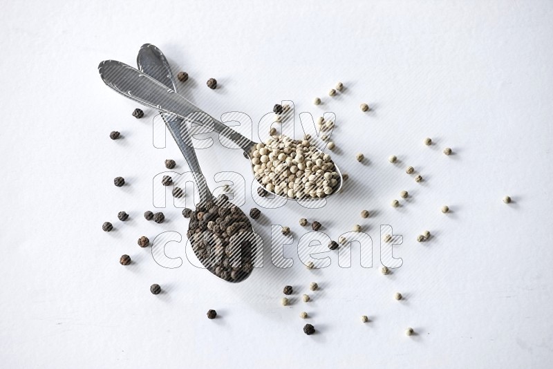 2 metal spoons full of black and white pepper beads with spreaded beads on white flooring