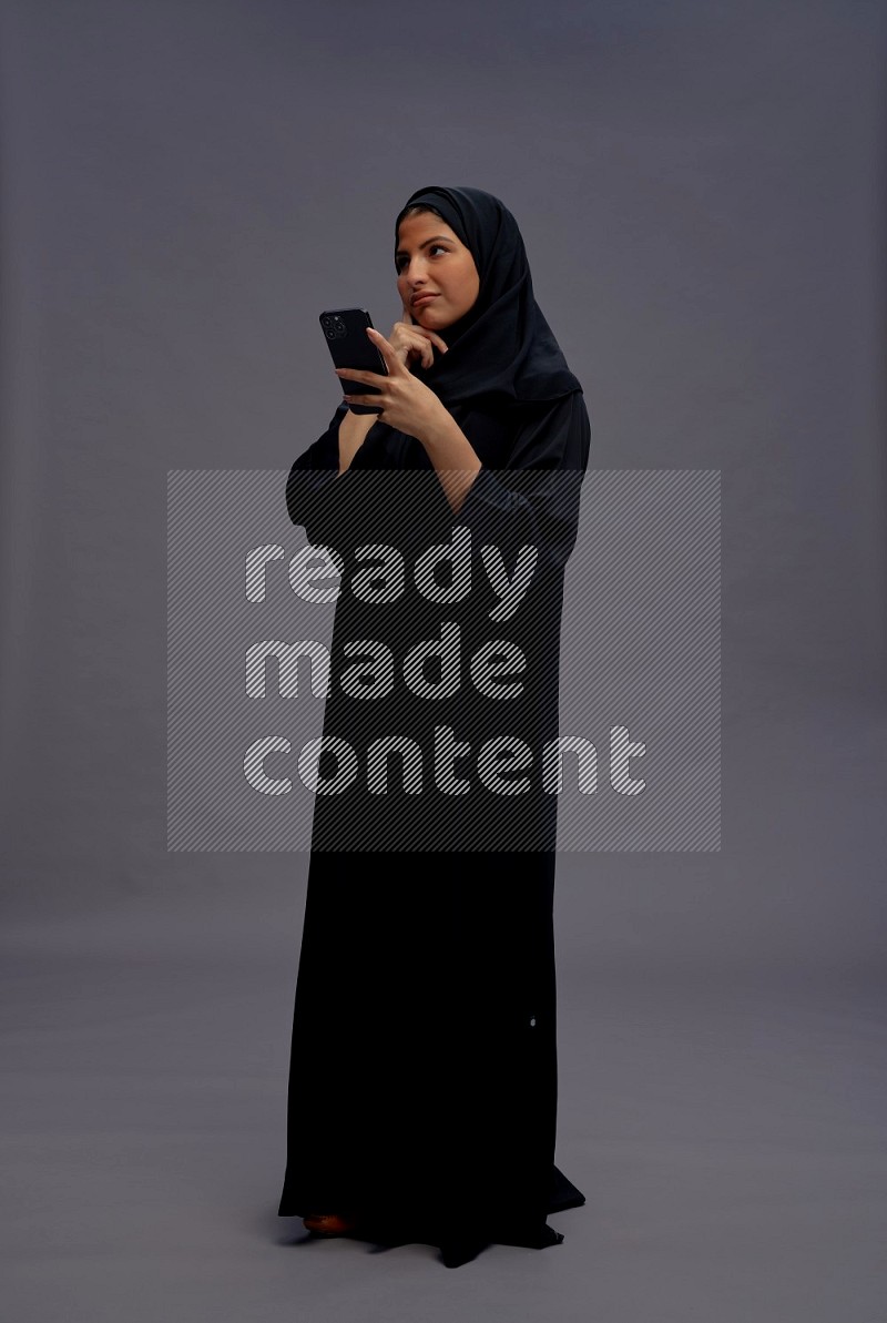 Saudi woman wearing Abaya standing texting on phone on gray background