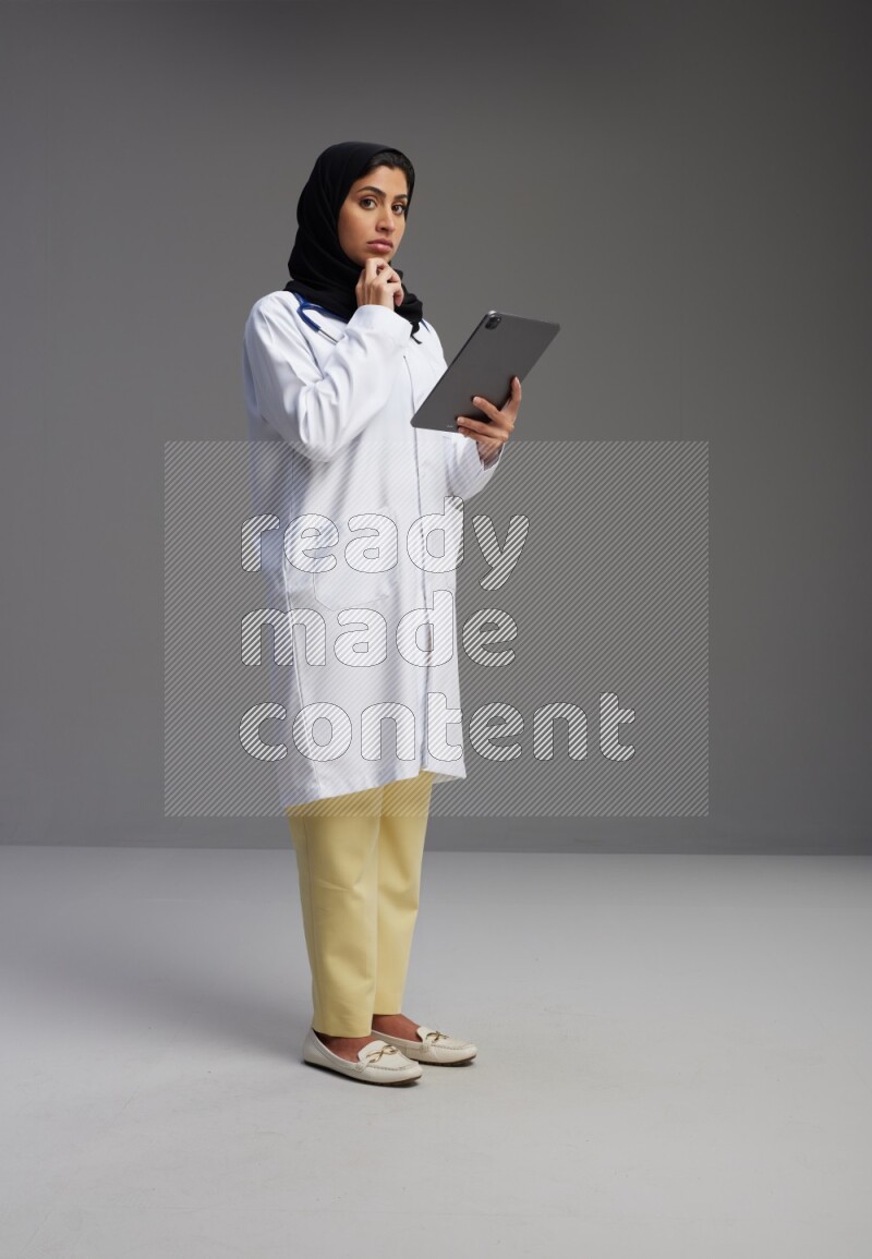Saudi woman wearing lab coat with stethoscope standing working on tablet on Gray background