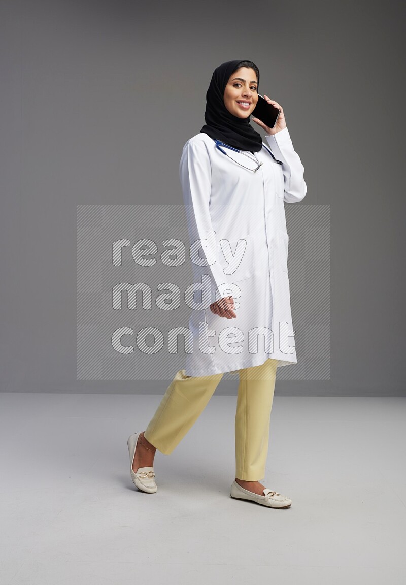 Saudi woman wearing lab coat with stethoscope standing talking on phone on Gray background