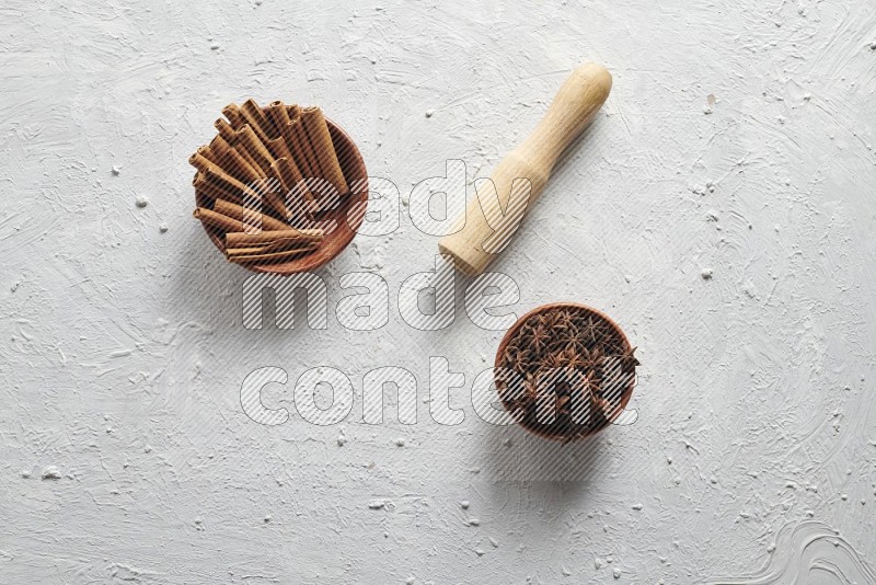 A wooden bowl full of cinnamon sticks and a wooden spoon with cinnamon powder are neatly placed beside a bowl of star anise on a textured white background
