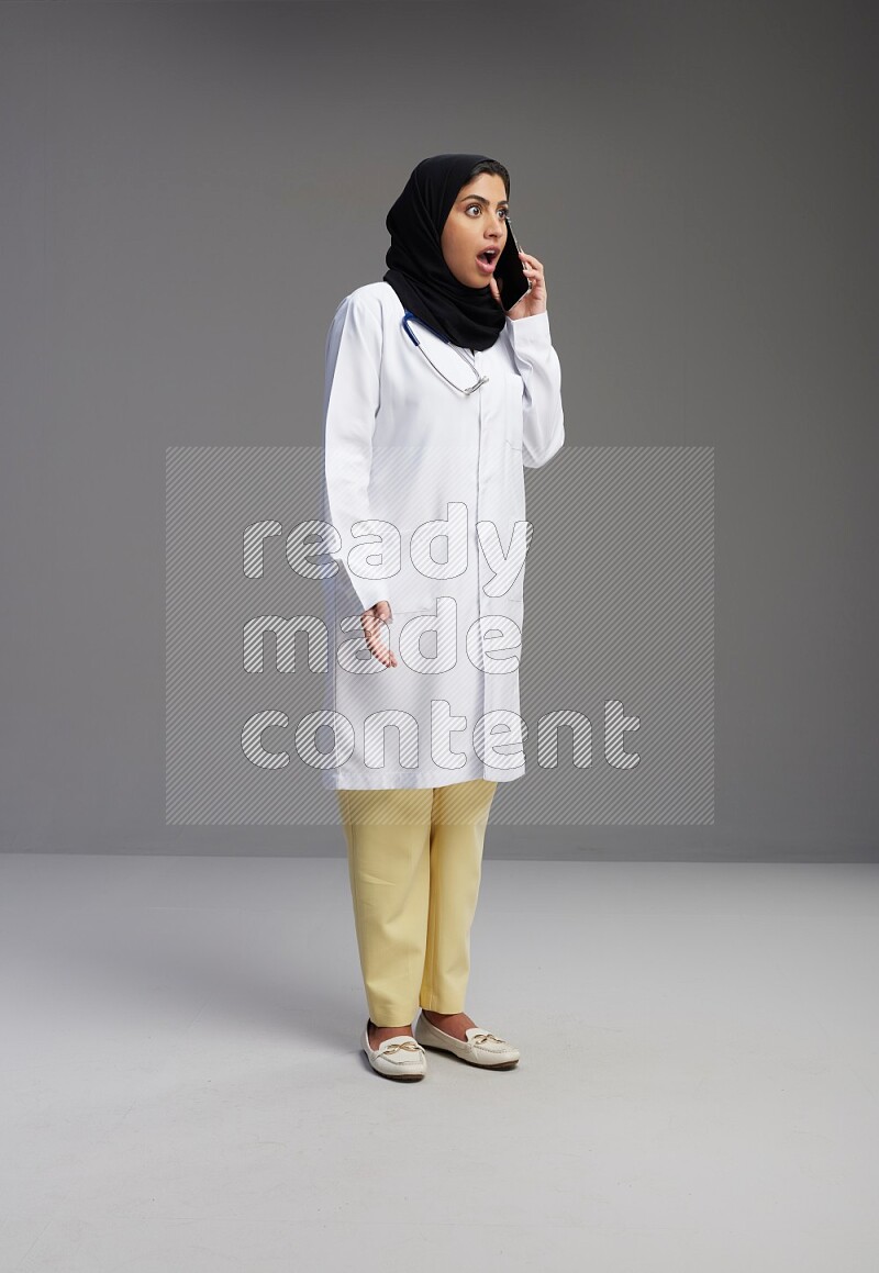 Saudi woman wearing lab coat with stethoscope standing talking on phone on Gray background