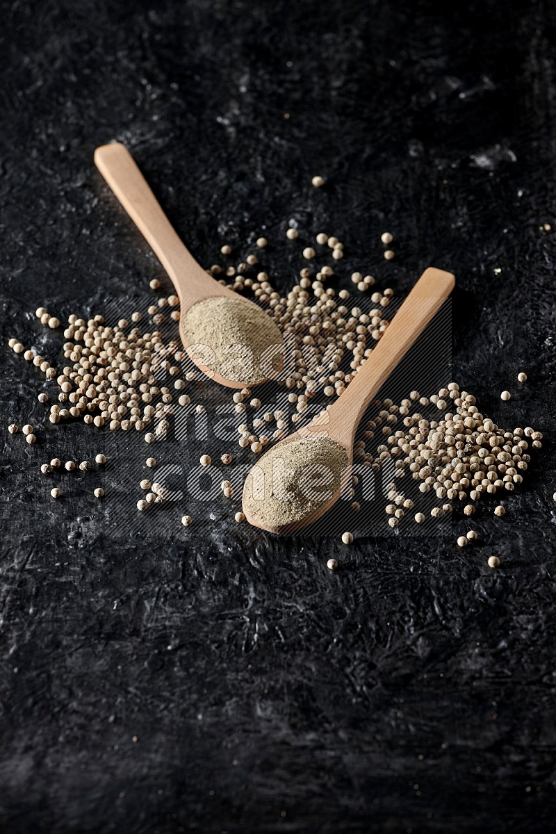 2 wooden spoons full of white pepper powder with white pepper beads on textured black flooring