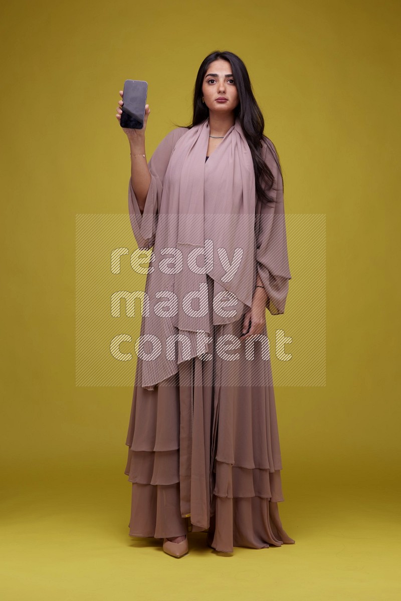 A woman Showing SmartPhone Screen on a Yellow Background wearing Brown Abaya