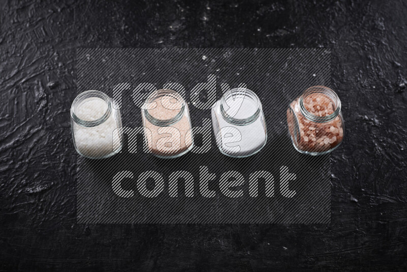 4 glass jars filled with table salt, coarse sea salt, fine himalayan salt and coarse himalayan salt on black background