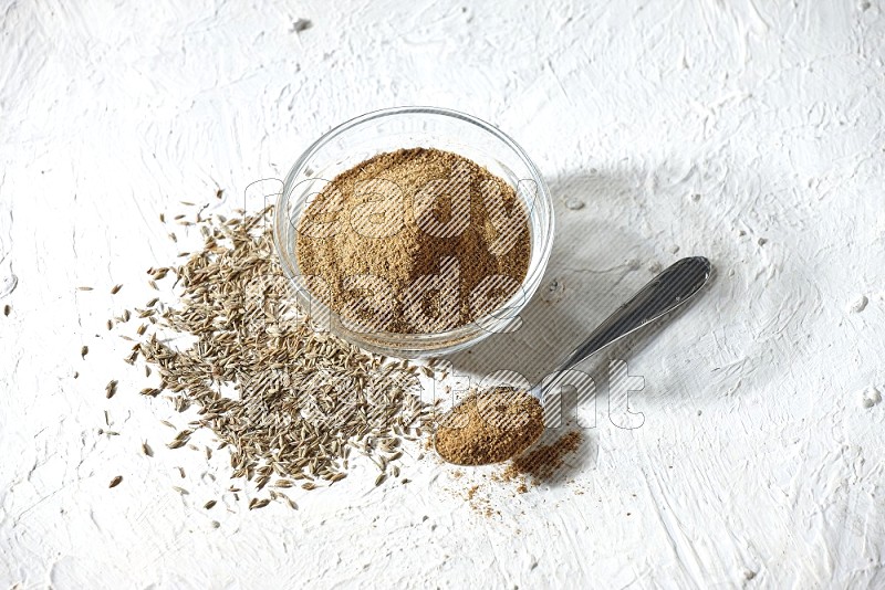 A glass bowl and metal spoon full of cumin powder and cumin seeds underneath it on textured white flooring