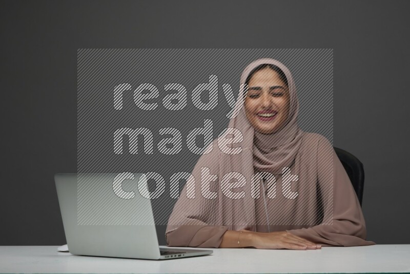 A Saudi woman Setting on her desk on a Gray Background wearing Brown Abaya with Hijab