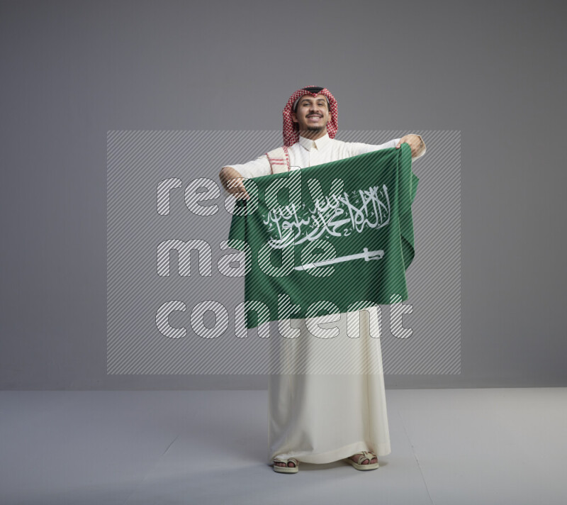 A saudi man standing wearing thob and red shomag holding big saudi flag on gray background