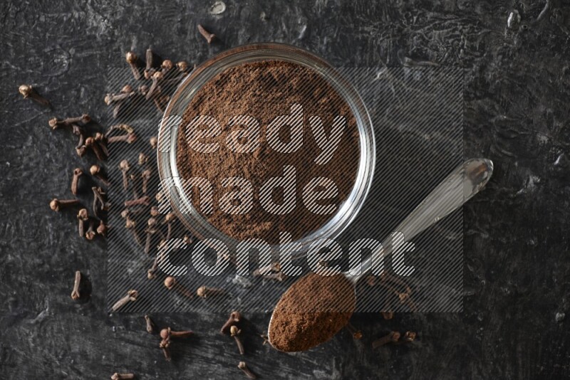 A glass bowl and a metal spoon full of cloves powder with gloves grains beside them on a textured black flooring