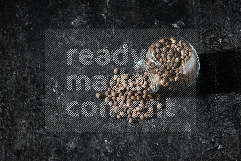 A flipped glass spice jar full of allspice whole balls and the balls spilled out of it on a textured black flooring