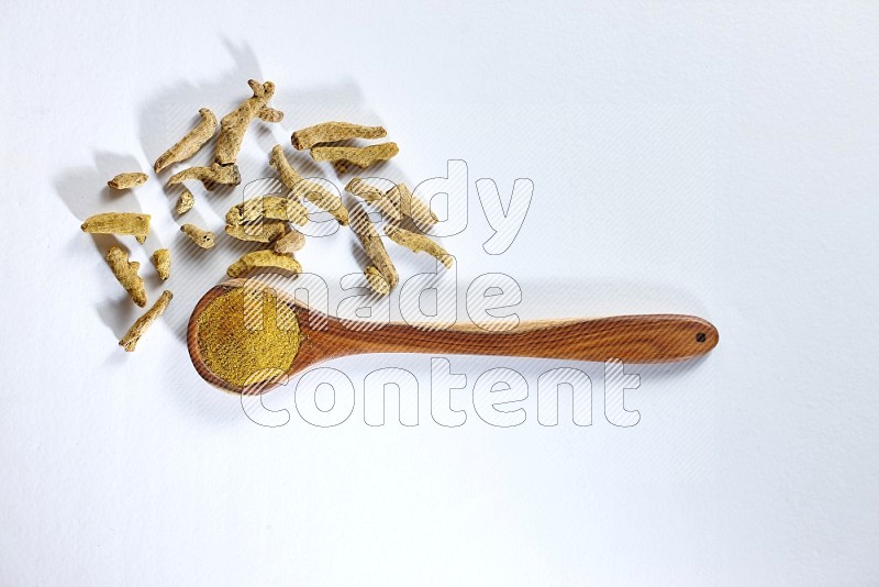 A wooden ladle full of turmeric powder and dried turmeric fingers beside it on white flooring