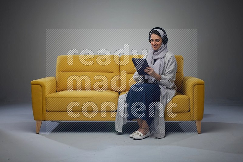 A Saudi woman wearing a light gray Abaya and head scarf sitting on a yellow sofa and using her phone while wearing headphones eye level on a grey background