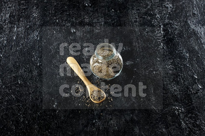A glass spice jar and a wooden spoon full of cumin seeds on a textured black flooring