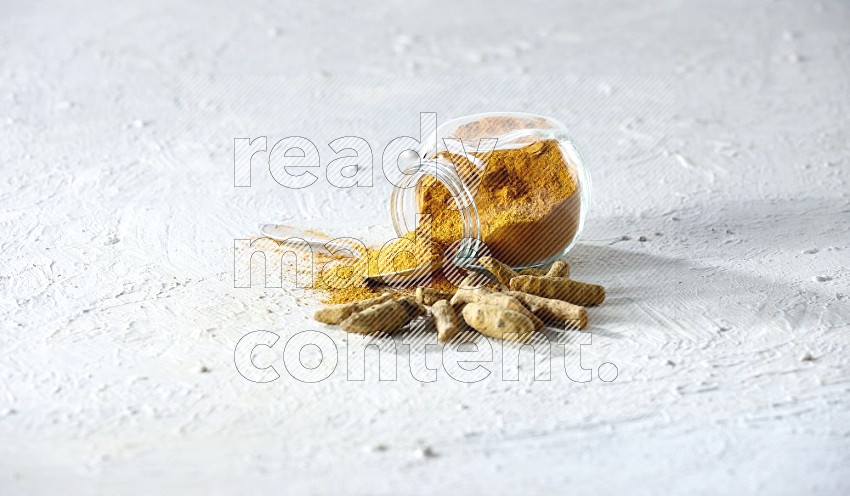 A glass spice jar and metal spoon full of turmeric powder and jar flipped and powder fell out with dried whole fingers on textured white flooring