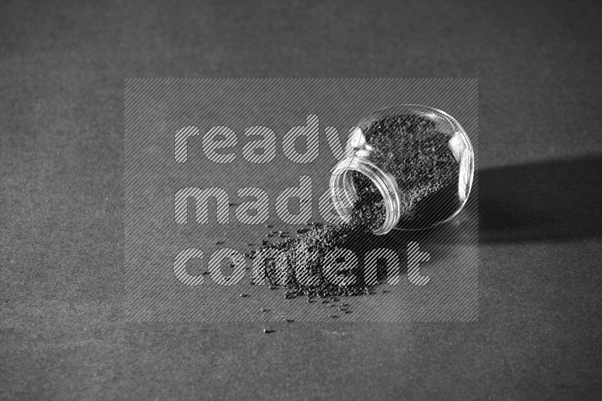 A glass spice jar full of black seeds flipped and seeds spreaded out on a black flooring in different angles