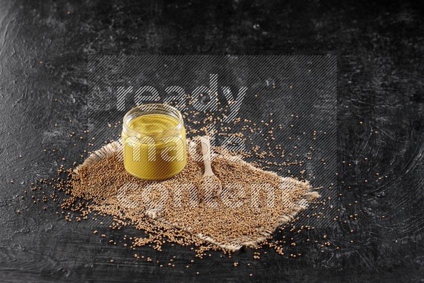 A glass jar full of mustard paste set on a burlap piece and a wooden spoon full of mustard seeds on a textured black flooring in different angles