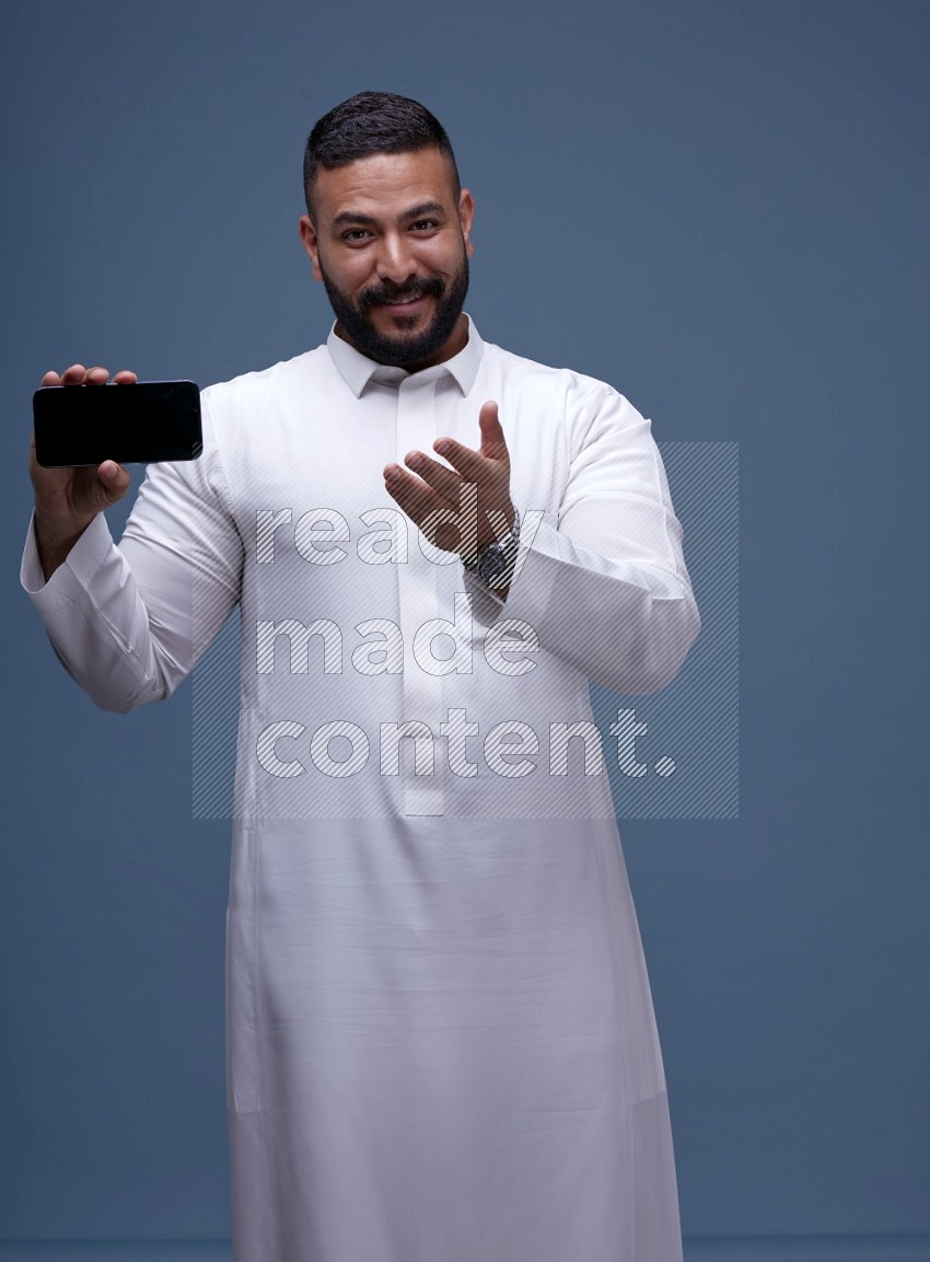 A man Showing his phone in Blue Background wearing Saudi Thob