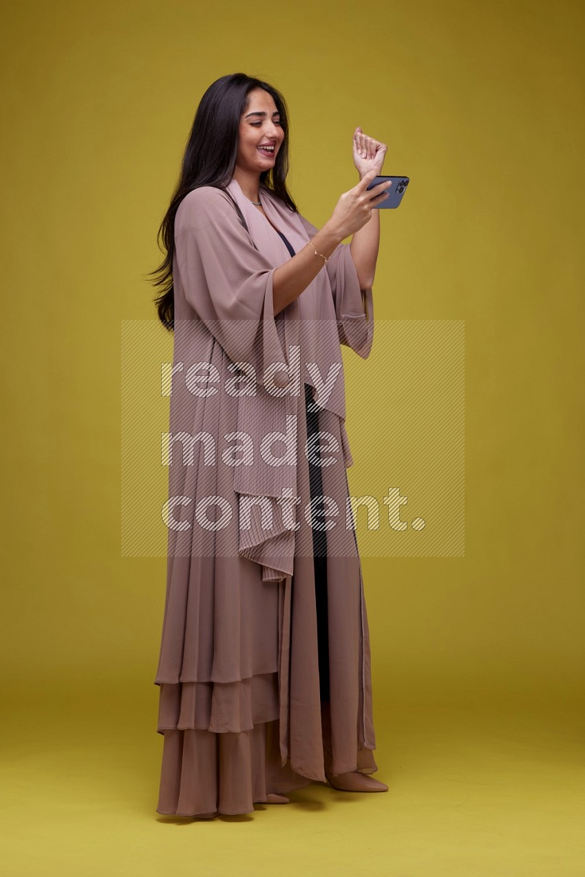 A woman Playing a Game on her smartphone on a Yellow Background wearing Brown Abaya