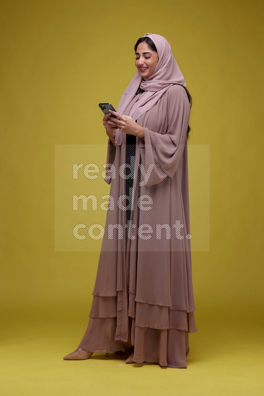 A woman Texting on her phone on  a Yellow Background wearing Brown Abaya with Hijab
