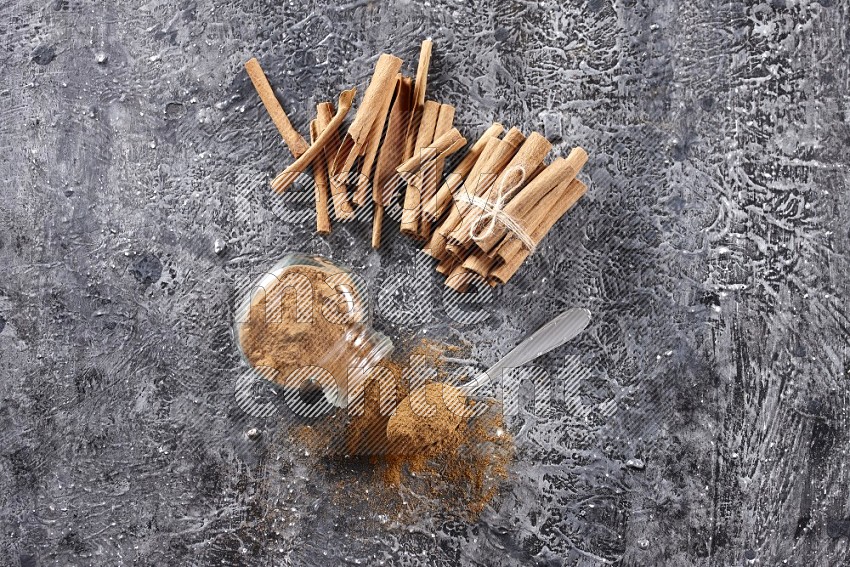 Herbal glass jar full cinnamon powder flipped and a metal spoon full of powder, cinnamon sticks stacked and bounded in the back on textured black background in different angles