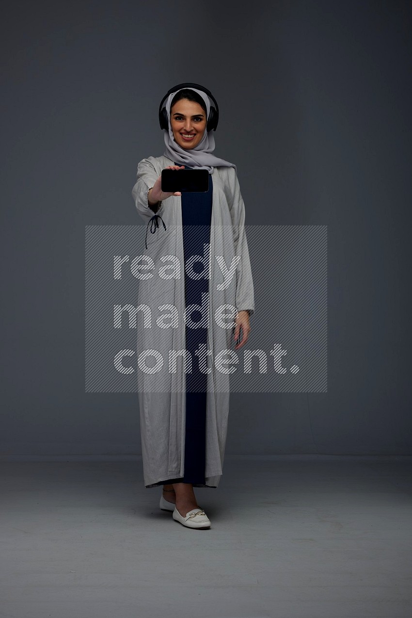 A Saudi woman wearing a light gray Abaya and head scarf setting on a dark grey chair and watching on the tablet eye level on a grey background