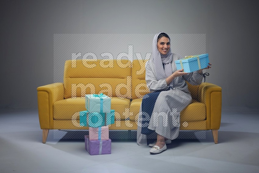 A Saudi woman wearing a light gray Abaya and head scarf sitting on a yellow sofa holding a gift box beside multi color gift boxes eye level on a grey background