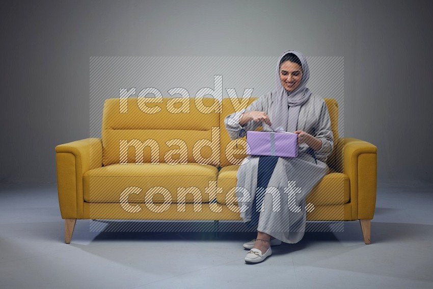 A Saudi woman wearing a light gray Abaya and head scarf sitting on a yellow sofa holding a gift box beside multi color gift boxes eye level on a grey background