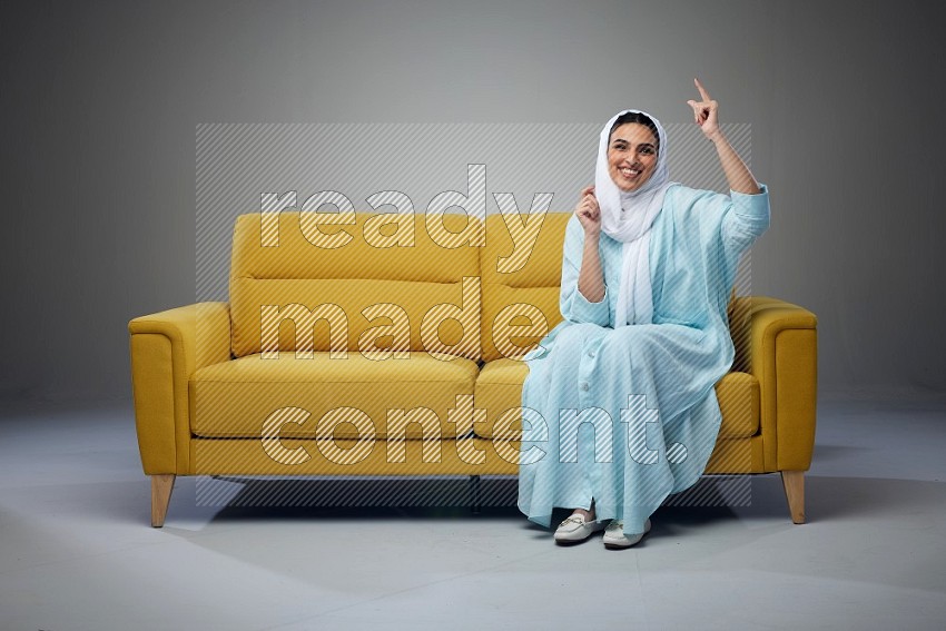 A Saudi woman wearing a light blue Abaya and white head scarf sitting on a yellow sofa and thinking and pointing up eye level on a grey background