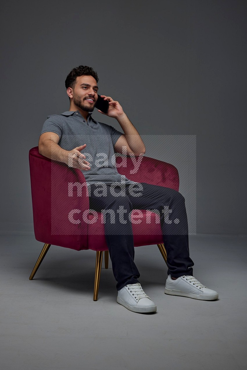 A man wearing casual and talking in his phone while setting on a burgundy chair eye level on a gray background