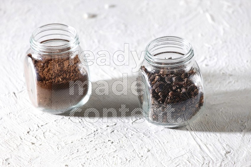 2 glass jars full of cloves powder and cloves grains on a textured white flooring