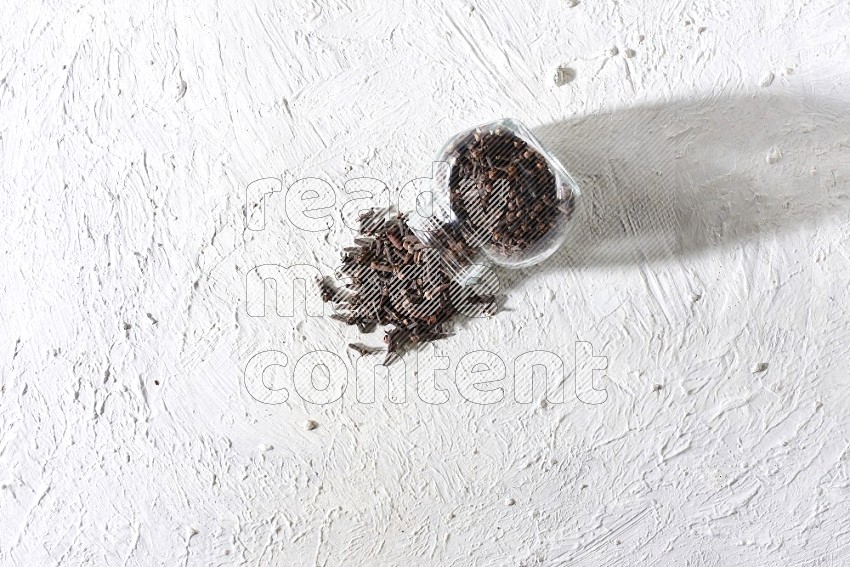 A glass spice jar full of cloves flipped and cloves came out of it on textured white flooring